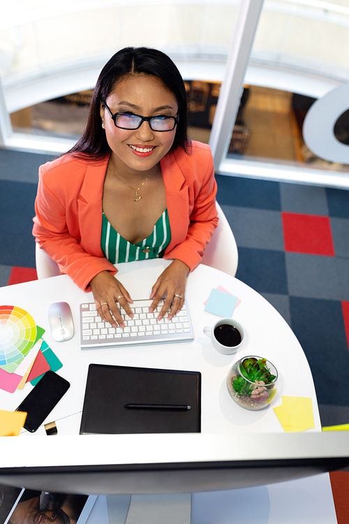 High angle view of pretty young Asian female Graphic designer working on computer at desk in office. This is a casual creative start-up business office for a diverse team