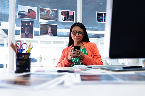 Front view of young pretty Asian female graphic designer using mobile phone at desk in office. This is a casual creative start-up business office for a diverse team