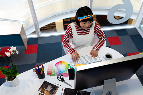 High angle view of young pretty Asian Female graphic designer working on computer at desk in office. This is a casual creative start-up business office for a diverse team