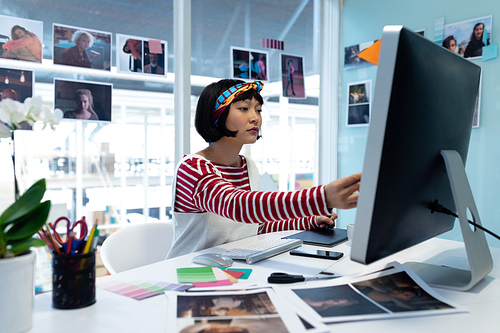 Front view of young pretty Asian female graphic designer using graphic tablet at desk in office. This is a casual creative start-up business office for a diverse team