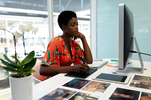 Side view of pretty young African american female graphic designer working on graphic tablet at desk in office. This is a casual creative start-up business office for a diverse team