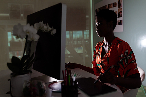 Side view of young pretty African-American female graphic designer using graphic tablet at desk in office. This is a casual creative start-up business office for a diverse team