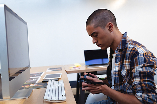 Side view of mixed race male graphic designer checking a digital camera at desk in office. This is a casual creative start-up business office for a diverse team