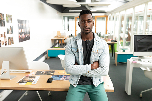 Portrait of African american male graphic designer sitting with arms crossed at desk in office. This is a casual creative start-up business office for a diverse team