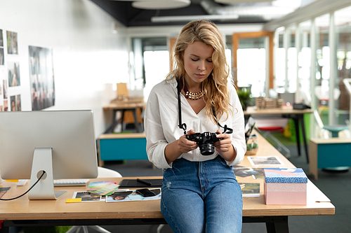 Front view of Caucasian female graphic designer reviewing photos on digital camera at desk in office. This is a casual creative start-up business office for a diverse team