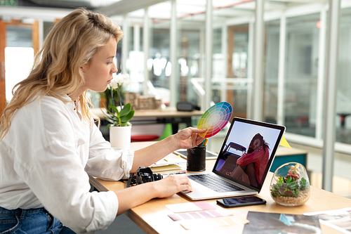 Side view of Caucasian female graphic designer looking at color swatch while using laptop at desk in office. This is a casual creative start-up business office for a diverse team