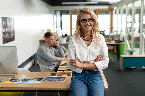 Portrait of Caucasian female graphic designer sitting with arms crossed at desk in office. Caucasian executive man working in the background. This is a casual creative start-up business office for a diverse team