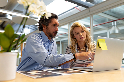 Side view of Caucasian male and female graphic designers discussing over laptop at desk in office. This is a casual creative start-up business office for a diverse team