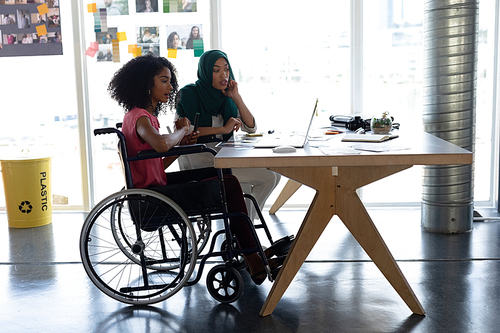 Side view of diverse female graphic designers discussing over laptop at desk in office. This is a casual creative start-up business office for a diverse team