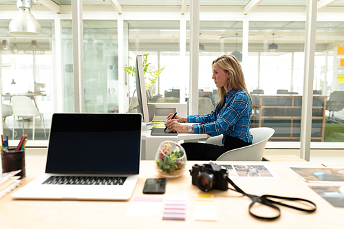 Side view of young Caucasian female graphic designer using graphic tablet at desk. This is a casual creative start-up business office for a diverse team