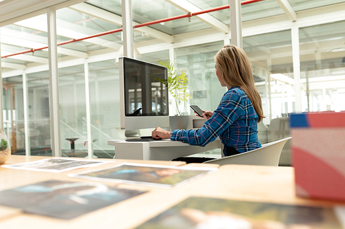 Rear view of Caucasian female graphic designer using mobile phone while working on graphic tablet at desk in office. This is a casual creative start-up business office for a diverse team