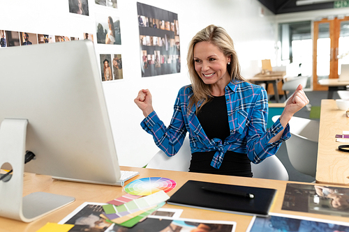 Front view of happy Caucasian female graphic designer celebrating success at desk in office. This is a casual creative start-up business office for a diverse team