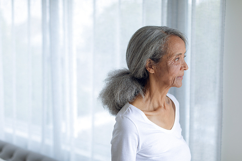 Side view of thoughtful senior mixed race woman standing and looking out the window. Authentic Senior Retired Life Concept