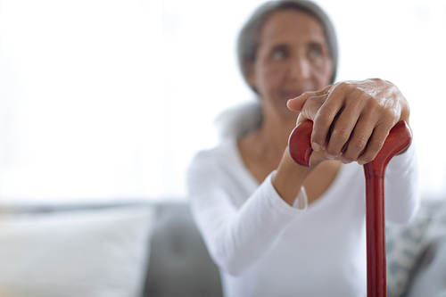 Side view of thoughtful senior mixed race woman sitting on couch while holding a brown walking stick. Authentic Senior Retired Life Concept