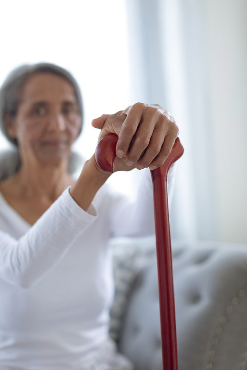 Side view of thoughtful senior mixed race woman sitting on couch while holding a brown walking stick. Authentic Senior Retired Life Concept