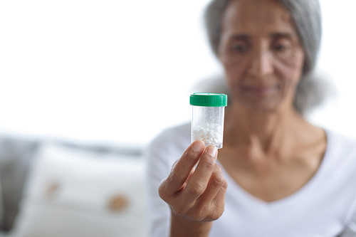 Side view of thoughtful senior mixed race woman sitting on couch while holding a small pill bottle. Authentic Senior Retired Life Concept