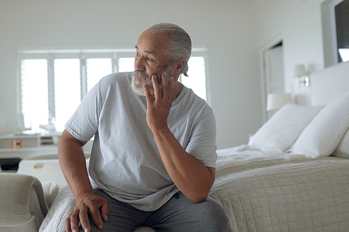 Front view of senior Caucasian man sitting on the bed in white bedroom. Authentic Senior Retired Life Concept