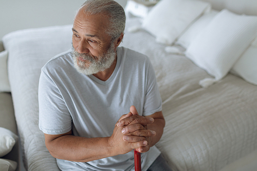 Side view of thoughtful senior Caucasian man sitting on the bed while holding a wooden walking stick in bedroom. Authentic Senior Retired Life Concept