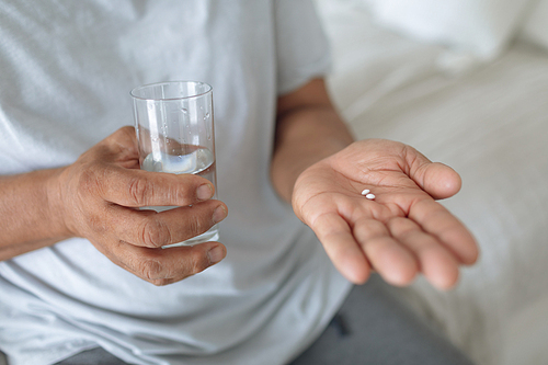 Close up of senior Caucasian man sitting on the bed while holding pills and a glass of water. Authentic Senior Retired Life Concept