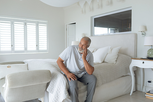 Side view of thoughtful senior Caucasian man sitting on the bed in bedroom. Authentic Senior Retired Life Concept