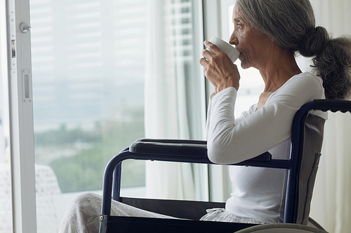 Side view close up of woman on wheelchair drinking coffee indoor. Authentic Senior Retired Life Concept