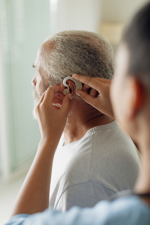 Rear view of healthcare worker putting hearing aid indoor. Authentic Senior Retired Life Concept