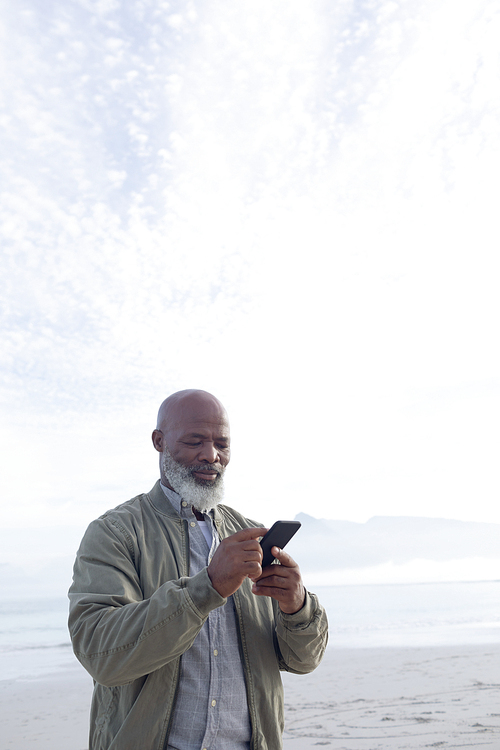 Low angle view of senior African-American man using mobile phone while standing on the beach on a beautiful day with mountains in the background. Authentic Senior Retired Life Concept