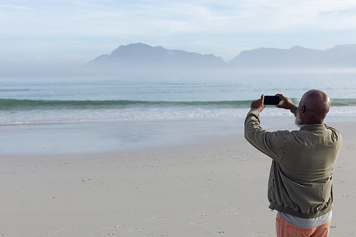 Rear view of handsome senior African-American man taking a picture of the sea and mountains on beach on beautiful day. Authentic Senior Retired Life Concept