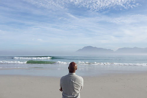 Rear view of handsome senior African-American standing with arms crossed and looking at the sea and mountains on beach on beautiful day. Authentic Senior Retired Life Concept