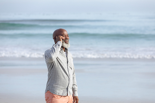 Front view of handsome senior African-American man talking on mobile phone while standing on the beach on beautiful day with mountains in the background. Authentic Senior Retired Life Concept