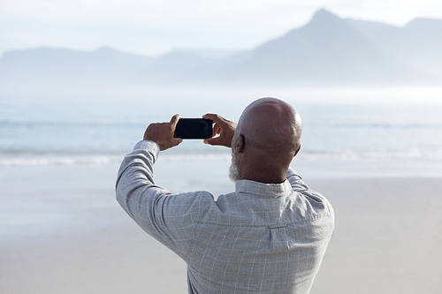 Rear view of handsome senior African-American man taking a picture of the sea and mountains on beach on beautiful day. Authentic Senior Retired Life Concept