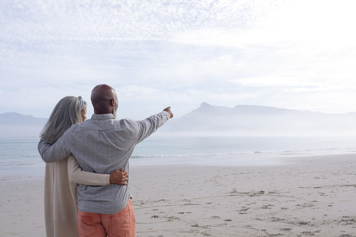 Rear view of happy senior African-American couple standing next to each other while looking at the sea and mountains on beach on beautiful cloudy day. African-American man points with right hand to the mountains. Authentic Senior Retired Life Concept