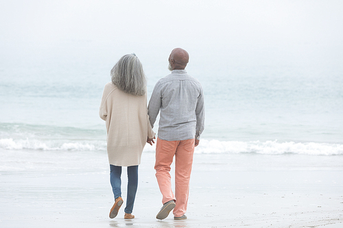 Rear view of happy senior African-American couple walking towards sea while holding hands on beach on cloudy day. Authentic Senior Retired Life Concept
