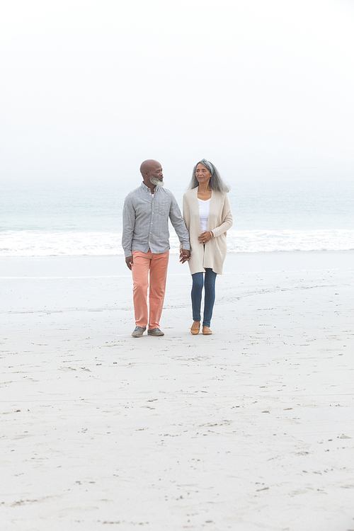 Front view of happy senior African-American couple walking away from the sea while holding hands on beach on cloudy day. Authentic Senior Retired Life Concept