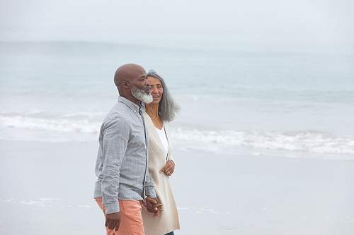 Side view of handsome senior African-American walking on the beach with his hands in pockets on beautiful day. Authentic Senior Retired Life Concept