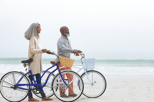 Side view of handsome senior African-American couple walking with bicycles on the beach with his hands in pockets on beautiful day. Authentic Senior Retired Life Concept