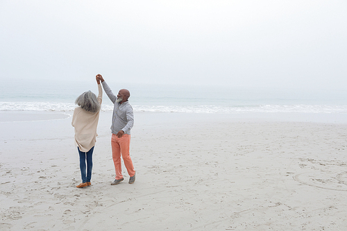 Front view of happy senior diverse couple dancing on the beach on cloudy day. Authentic Senior Retired Life Concept