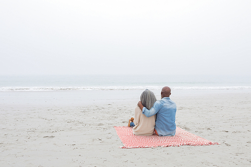 Rear view of diverse Couple sitting on red and white striped picnic blanket at the beach. Authentic Senior Retired Life Concept