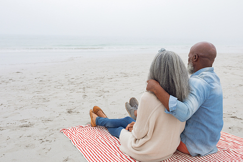 Rear view of happy senior diverse couple sitting on red and white striped picnic blanket at the beach. Authentic Senior Retired Life Concept