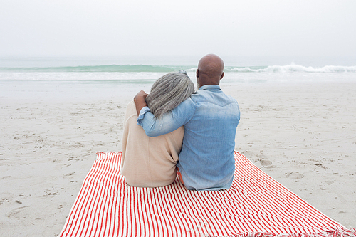 Rear view of happy senior diverse couple sitting on red and white striped picnic blanket at the beach. Authentic Senior Retired Life Concept