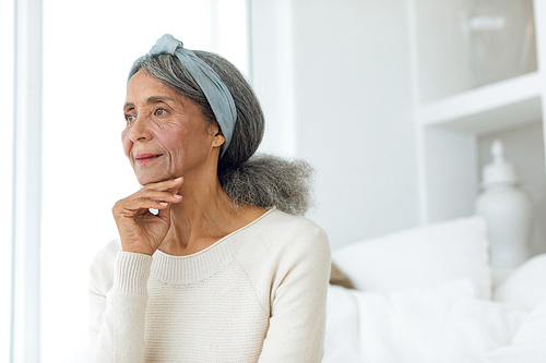 Front view of happy senior mixed race woman thinking while sitting on white couch. Authentic Senior Retired Life Concept