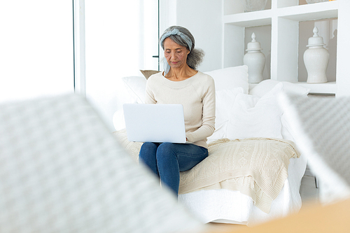 Front view of beautiful senior mixed race woman sitting on couch while using a white laptop in beach house. Authentic Senior Retired Life Concept
