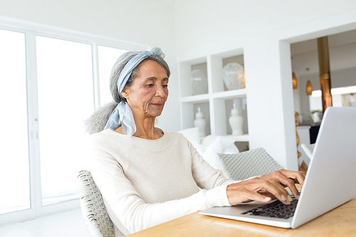Side view of senior mixed race woman using a white laptop on a table in beach house. Authentic Senior Retired Life Concept