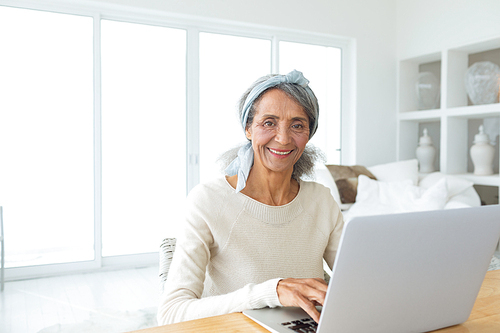 Side view of senior mixed race woman using a white laptop on a table in beach house. Authentic Senior Retired Life Concept