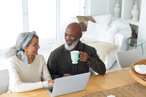 Front view of diverse senior couple using laptop on table while man holds a cup in beach house. Authentic Senior Retired Life Concept