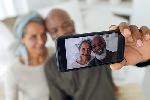 Front view of diverse senior couple sitting on a couch inside a room while taking picture with smartphone in beach house. Authentic Senior Retired Life Concept