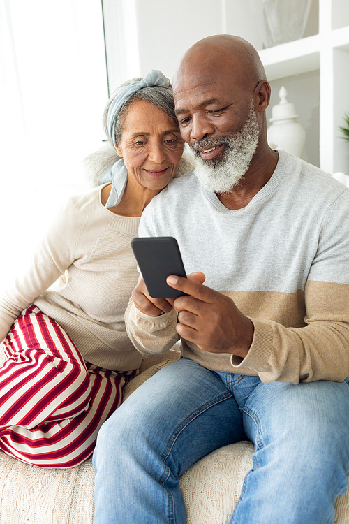Front view of happy diverse senior couple using a smartphone on sofa in beach house. Authentic Senior Retired Life Concept