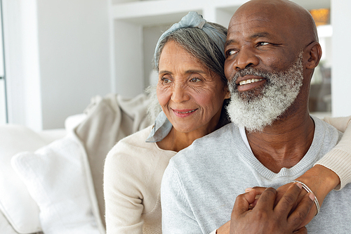 Front view of happy senior diverse couple sitting on white couch in beautiful beach house. Authentic Senior Retired Life Concept