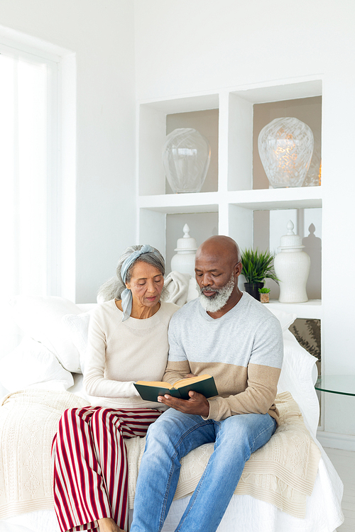 Front view of happy senior diverse couple reading a book while sitting on white sofa in beach house. Authentic Senior Retired Life Concept