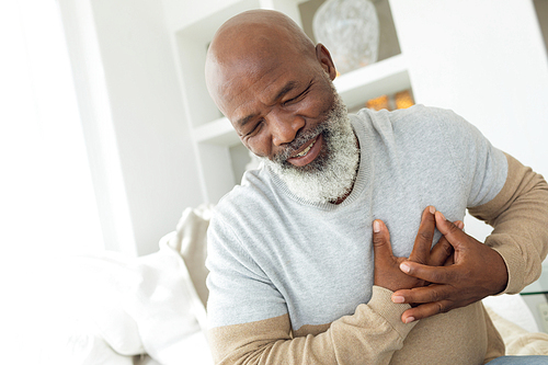 Front view of handsome senior African American man smiling while sitting on white sofa in beach house. He is touching his chest. Authentic Senior Retired Life Concept